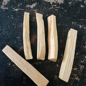 May include: Five pieces of light brown palo santo wood sticks on a dark gray textured surface.