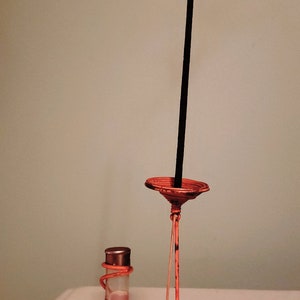 Pu&ograve; includere: Una bottiglia di vetro trasparente con un'etichetta che recita "Vantine Incense Lilac" &egrave; appoggiata su un supporto in metallo arancione. Un bastoncino di incenso nero &egrave; nel supporto.