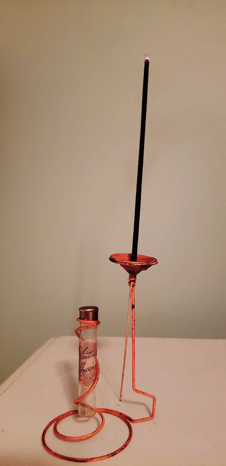 May include: A clear glass bottle with a label that reads "Vantine Incense Lilac" is sitting on an orange metal stand. A black incense stick is in the stand.