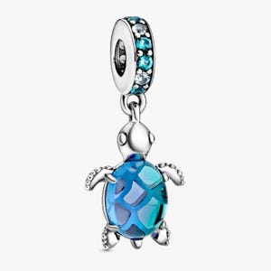 Op de afbeelding: Een zilveren bedel met een zeeschildpadontwerp. De schildpad heeft een blauw en blauwgroen schild met een geometrisch patroon. De bovenste ring van de bedel is versierd met blauwe en heldere edelstenen. Een sieraad.