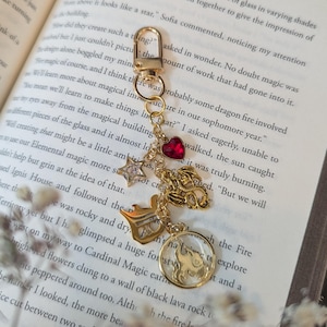 May include: Gold-colored keychain with various charms, including a star, heart, and a circular pendant with a design. The keychain is displayed on an open book with text and a soft, neutral background.