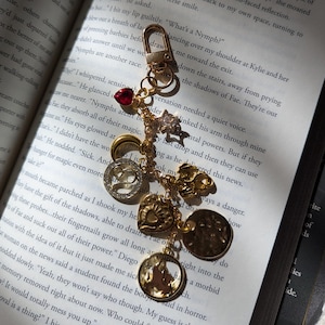 May include: Gold-toned charm bracelet with various charms, including a heart, a star, and a round charm with a silhouette. The bracelet is attached to a clasp. The background is an open book.