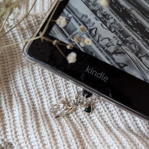 May include: A Kindle e-reader with a clear protective case. A silver charm with a deer design and a green heart-shaped stone hangs from the device. The word "kindle" is visible on the device. The e-reader rests on a white knitted surface.