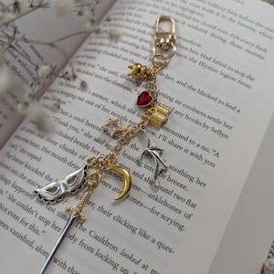 May include: A gold-toned keychain with various charms, including a red heart, a crescent moon, a sword, and a masquerade mask. The keychain is displayed on an open book with text visible.