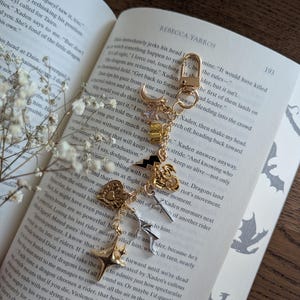 May include: Gold-toned keychain with various charms, including a star, lightning bolt, and heart, resting on an open book. The book's pages display text and decorative dragon illustrations. The keychain is a decorative accessory.