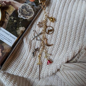 May include: Gold-coloured keychain featuring charms like stars, a bow and arrow, a moon, a sword, and a red rose. The keychain is attached to a clear phone case with decorative stickers. The background is a cream-coloured knitted fabric.