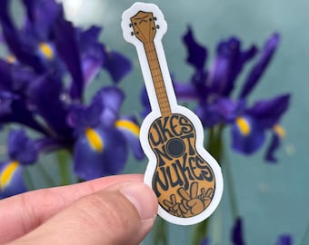 Op de afbeelding: Een sticker in de vorm van een ukelele met de woorden "Ukes Not Nukes" in bruine letters. De sticker heeft ook een vredesteken. De achtergrond omvat paarse bloemen en een turquoise muur.