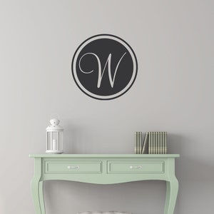 Op de afbeelding: Een muursticker met een zwarte cirkel en de witte letter "W" in sierlijke letters. Het is bevestigd op een lichtgrijze muur boven een mintgroene console, een witte poef en een witte lantaarn.