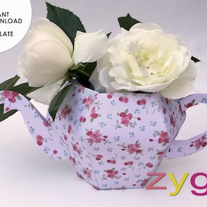 May include: A white paper teapot with a floral pattern, shaped like a teapot, with a handle and spout. The teapot is filled with white roses.