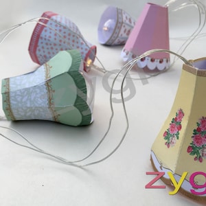 May include: A string of fairy lights with paper lampshades. The lampshades are decorated with floral patterns in pink, green, and yellow.
