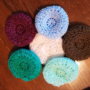 May include: A collection of six handmade, circular crochet items in various colors: deep burgundy, light blue, white, brown, teal, and light turquoise. The items are arranged in a circular pattern on a wooden surface. These could be used as coasters or decorative items.