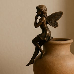 Könnte beinhalten: Eine bronzefarbene Figur mit Flügeln, die auf dem Rand einer beigen Vase sitzt. Die Figur hat kurzes Haar, ein kurzärmeliges Oberteil und einen Rock, eine Hand ist zum Kinn geführt und die Beine hängen über den Rand der Vase hinunter.