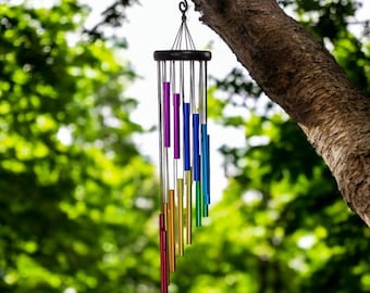 Puede incluir: Un carillón de viento con una parte superior circular de madera oscura y múltiples tubos metálicos colgantes en un vibrante degradado de arcoíris de rojo, naranja, amarillo, verde, azul y morado. El carillón cuelga de un gancho de metal negro.