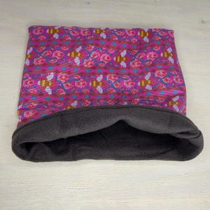 May include: A reversible cowl scarf with a vibrant pink floral pattern on one side and a dark gray fleece lining on the other. The scarf is folded, showcasing the contrasting textures and colors.