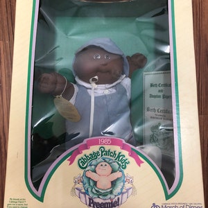 Op de afbeelding: Een Cabbage Patch Kids Preemie pop in een blauw-wit outfit. De pop heeft een bruine huid en zwart haar. De pop is verpakt in een doos met een groene achtergrond en de tekst "1985 Cabbage Patch Kids Preemie!" De doos bevat ook de tekst "Ik ben uniek. Ik ben adopteerbaar!" en "Leeftijd 3 jaar en ouder".