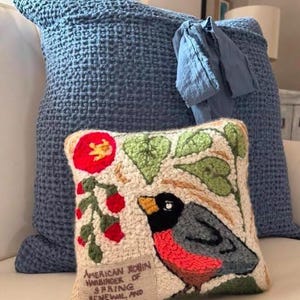 May include: A cream-colored pillow with an embroidered image of a red-breasted robin perched on a branch with green leaves and red berries. The pillow has the text "American Robin Harbinger of Spring Renewal and New Beginnings" embroidered on the bottom.