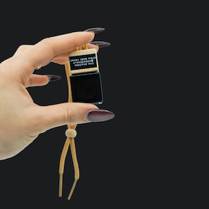 Op de afbeelding: Een zwarte glazen autodiffuser met een houten bovenkant en een beige touwtje. De diffuser heeft de tekst "CAR DIFFUSER  GOODIE SCENTS  GOODIE MARK  MORE" gedrukt op de houten bovenkant.