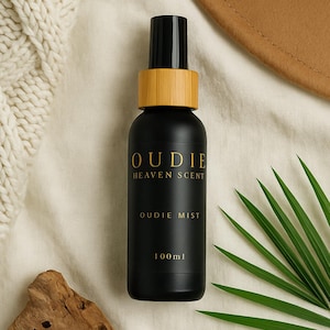 May include: A black Oudie Heaven Scent mist bottle with gold lettering, a bamboo collar, and a black cap. The bottle is labeled "Oudie Mist" and "100ml". The background includes a white knitted blanket, a brown hat, a piece of driftwood, and a green palm leaf.
