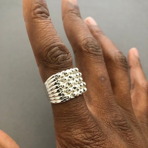 Puede incluir: Un anillo plateado con un dise&ntilde;o texturizado que presenta peque&ntilde;os elementos redondos y elevados. El anillo se lleva en un dedo, mostrando sus intrincados detalles y su superficie reflectante. El dise&ntilde;o del anillo es un grupo de peque&ntilde;as esferas.