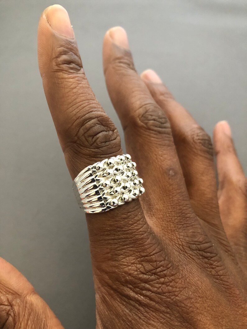 Puede incluir: Un anillo plateado con un dise&ntilde;o texturizado que presenta peque&ntilde;os elementos redondos y elevados. El anillo se lleva en un dedo, mostrando sus intrincados detalles y su superficie reflectante. El dise&ntilde;o del anillo es un grupo de peque&ntilde;as esferas.