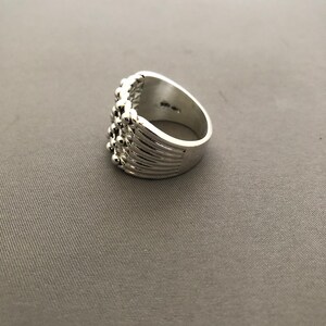 Puede incluir: Un anillo plateado con un dise&ntilde;o texturizado. El anillo presenta una serie de peque&ntilde;as cuentas redondas a lo largo de la parte superior y los lados. La banda tiene l&iacute;neas verticales. El anillo est&aacute; sobre un fondo gris.