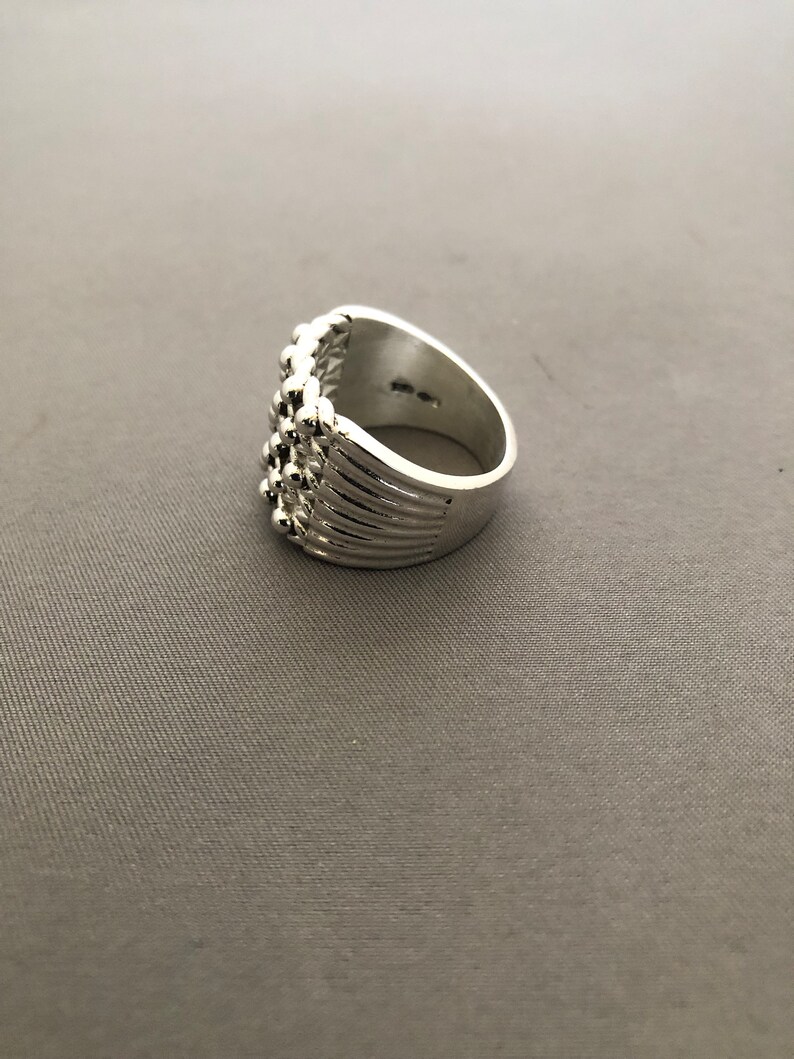 Puede incluir: Un anillo plateado con un dise&ntilde;o texturizado. El anillo presenta una serie de peque&ntilde;as cuentas redondas a lo largo de la parte superior y los lados. La banda tiene l&iacute;neas verticales. El anillo est&aacute; sobre un fondo gris.
