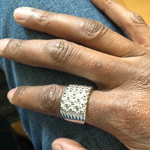 Puede incluir: Un anillo plateado con una superficie texturizada, usado en un dedo. El anillo tiene una banda ancha con un patr&oacute;n repetido de peque&ntilde;as formas redondeadas. El anillo se muestra sobre un fondo de mezclilla azul.