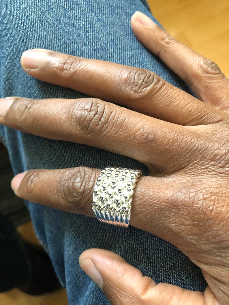 Puede incluir: Un anillo plateado con una superficie texturizada, usado en un dedo. El anillo tiene una banda ancha con un patr&oacute;n repetido de peque&ntilde;as formas redondeadas. El anillo se muestra sobre un fondo de mezclilla azul.
