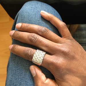 Puede incluir: Un anillo plateado con superficie texturizada, usado en una mano. El anillo tiene forma rectangular y est&aacute; cubierto de peque&ntilde;os detalles en relieve. La mano descansa sobre unos vaqueros azules.