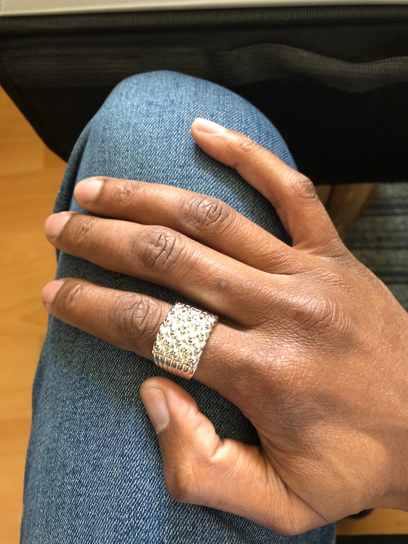 Puede incluir: Un anillo plateado con superficie texturizada, usado en una mano. El anillo tiene forma rectangular y est&aacute; cubierto de peque&ntilde;os detalles en relieve. La mano descansa sobre unos vaqueros azules.