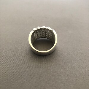 Puede incluir: Un anillo plateado con un dise&ntilde;o tejido alrededor de la banda. El anillo tiene un acabado pulido y una banda ancha. El patr&oacute;n tejido a&ntilde;ade textura e inter&eacute;s visual a la joya.
