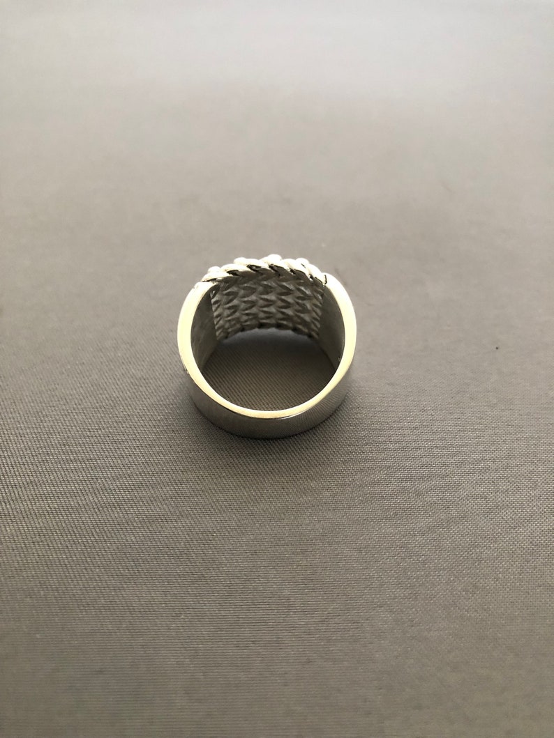 Puede incluir: Un anillo plateado con un dise&ntilde;o tejido alrededor de la banda. El anillo tiene un acabado pulido y una banda ancha. El patr&oacute;n tejido a&ntilde;ade textura e inter&eacute;s visual a la joya.