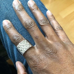 Puede incluir: Un anillo plateado con superficie texturizada. El anillo es ancho y rectangular, y se lleva en un dedo. El anillo se encuentra sobre un fondo de vaqueros azules.