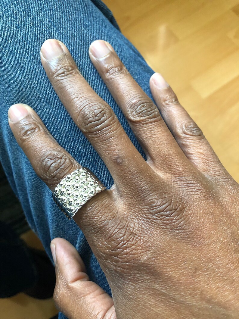 Puede incluir: Un anillo plateado con superficie texturizada. El anillo es ancho y rectangular, y se lleva en un dedo. El anillo se encuentra sobre un fondo de vaqueros azules.