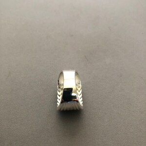 Puede incluir: Un anillo plateado con una banda ancha y un dise&ntilde;o texturizado en los laterales. El anillo tiene una superficie pulida y reflectante. Los lados del anillo presentan un patr&oacute;n repetido de peque&ntilde;as hojas o plumas estilizadas.