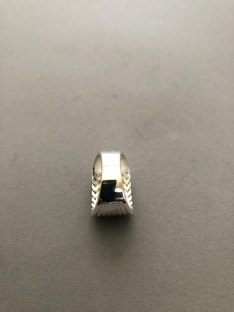 Puede incluir: Un anillo plateado con una banda ancha y un dise&ntilde;o texturizado en los laterales. El anillo tiene una superficie pulida y reflectante. Los lados del anillo presentan un patr&oacute;n repetido de peque&ntilde;as hojas o plumas estilizadas.
