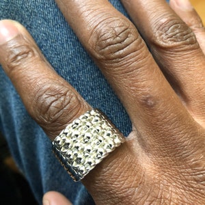 May include: A silver-colored ring with a textured surface, worn on a finger. The ring has a rectangular shape with a pattern of small, raised bumps. The ring is shown against a background of blue denim.