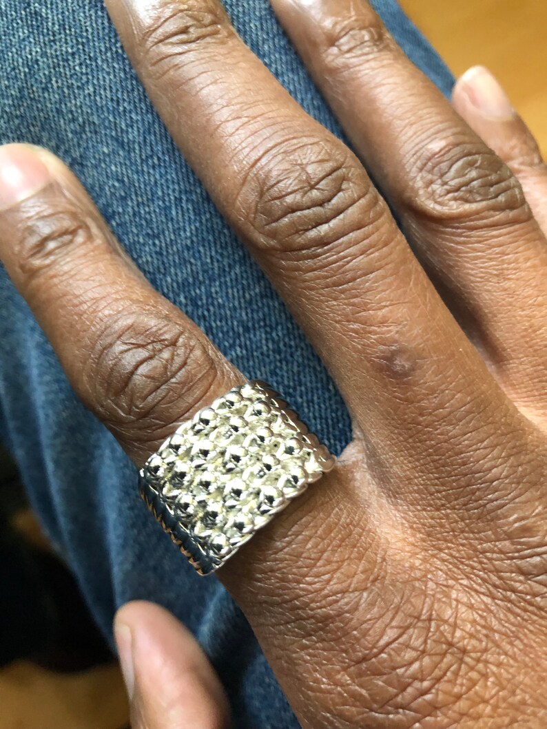 Puede incluir: Un anillo plateado con una superficie texturizada, usado en un dedo. El anillo tiene forma rectangular con un patr&oacute;n de peque&ntilde;as protuberancias en relieve. El anillo se muestra sobre un fondo de mezclilla azul.