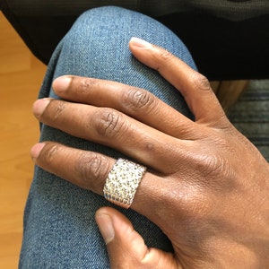 Puede incluir: Un anillo plateado con superficie texturizada se lleva en una mano. El anillo tiene una banda ancha y un patr&oacute;n de peque&ntilde;os detalles en relieve. La mano descansa sobre una pierna vestida con mezclilla.