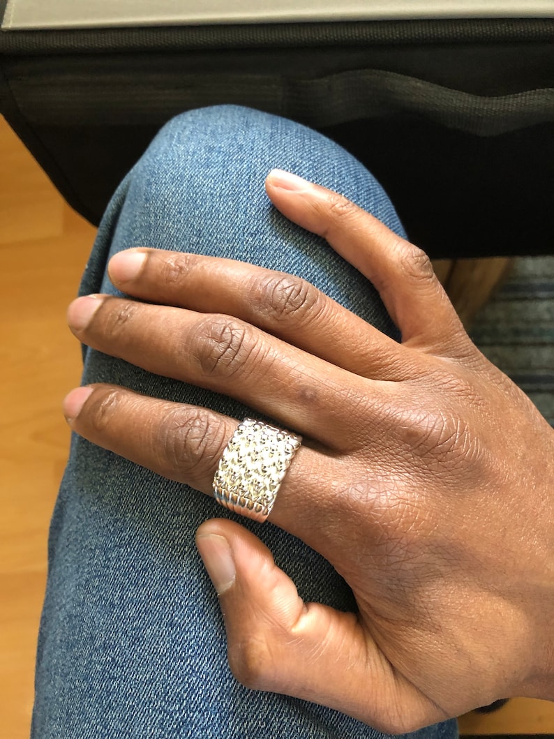 Puede incluir: Un anillo plateado con superficie texturizada se lleva en una mano. El anillo tiene una banda ancha y un patr&oacute;n de peque&ntilde;os detalles en relieve. La mano descansa sobre una pierna vestida con mezclilla.