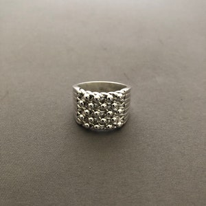 Puede incluir: Un anillo plateado con un dise&ntilde;o texturizado. El anillo presenta una banda con l&iacute;neas horizontales y una secci&oacute;n superior cubierta de peque&ntilde;as cuentas redondas en relieve. El anillo est&aacute; sobre un fondo gris.