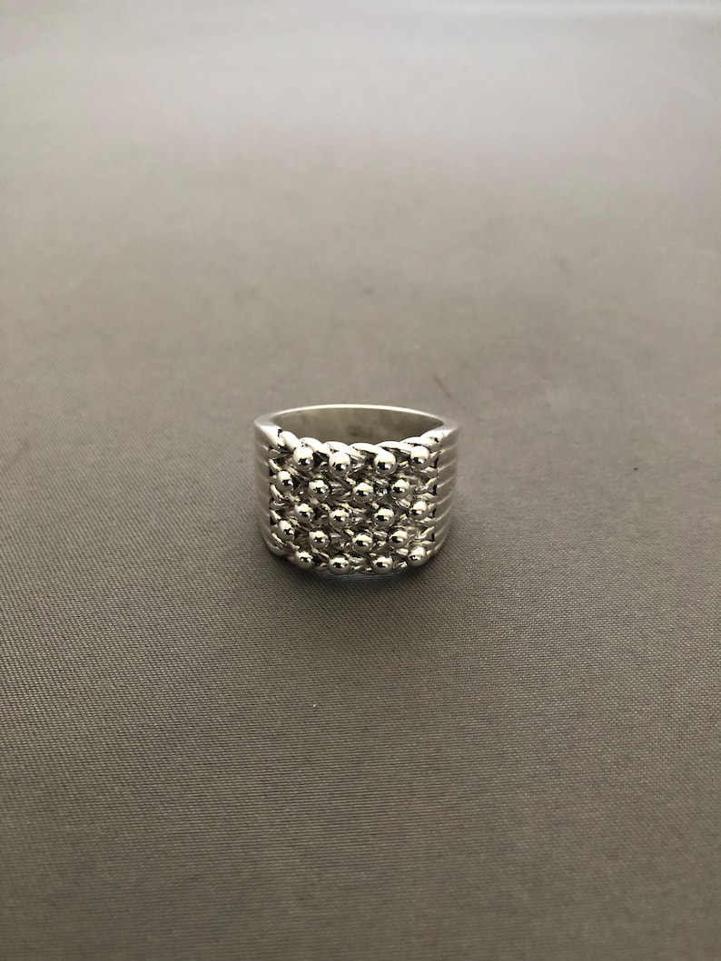 Puede incluir: Un anillo plateado con un dise&ntilde;o texturizado. El anillo presenta una banda con l&iacute;neas horizontales y una secci&oacute;n superior cubierta de peque&ntilde;as cuentas redondas en relieve. El anillo est&aacute; sobre un fondo gris.