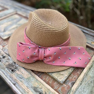 May include: A straw fedora hat with a pink band and a matching bow tie. The pink fabric features a repeating pattern of small white and black bird illustrations with yellow accents. The hat is set against a weathered wooden surface.