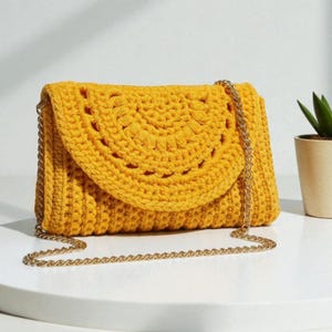 Könnte beinhalten: Senfgelbe Häkeltasche mit goldener Kette. Der Umschlag hat ein dekoratives, halbkreisförmiges Lochmuster. Die Tasche ist aus dickem, strukturiertem Garn gefertigt und liegt auf einer weißen Oberfläche.
