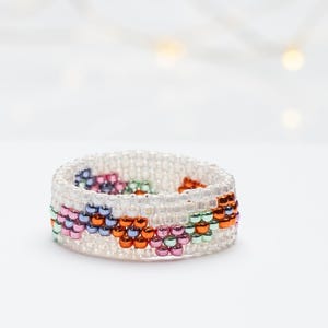 Op de afbeelding: Een kralenring gemaakt van kleine, doorzichtige kralen die een band vormen. De ring heeft een herhalend patroon van kleine bloemen gemaakt met kralen met een metallic afwerking in tinten roze, oranje, groen en paars op de doorzichtige basis.