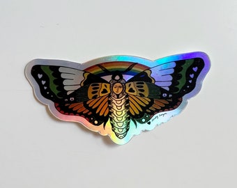 May include: Holographic sticker with a moth design and a rainbow. The moth is depicted with a face and is coloured in orange, yellow, green, and black. The sticker has a shiny, iridescent finish. The sticker is a decorative item.