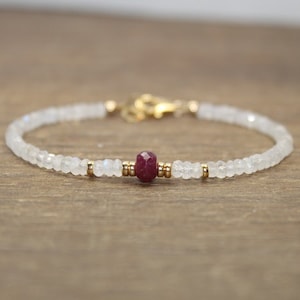 Puede incluir: Un delicado brazalete hecho con cuentas de piedra lunar blanca y un solo acento de piedra roja. El brazalete está encordado en alambre dorado.