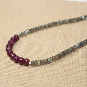 May include: A beaded necklace featuring a strand of faceted grey labradorite beads with iridescent blue flashes, accented by a section of deep red faceted ruby beads. The necklace is finished with small silver spacers between the gemstone beads.