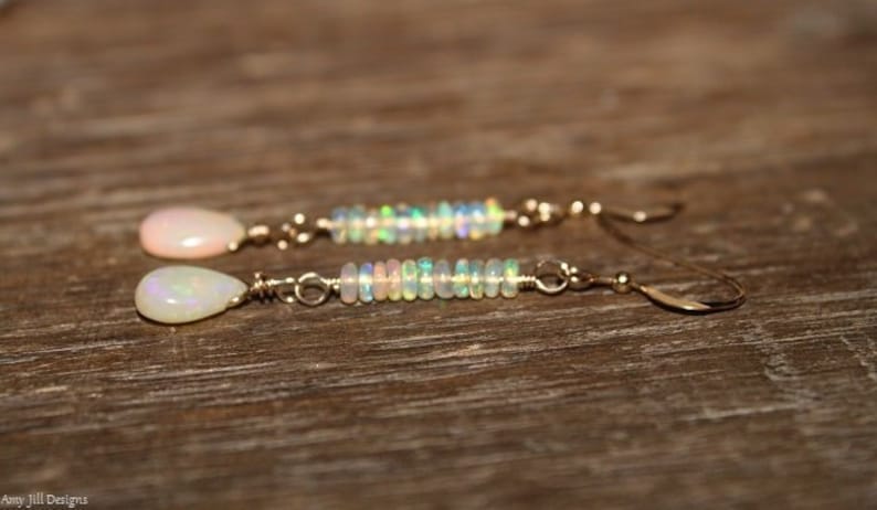 Puede incluir: Un par de pendientes dorados con una &oacute;palo en forma de l&aacute;grima y un dise&ntilde;o de &oacute;palo con cuentas. Los pendientes se muestran sobre una superficie de madera.