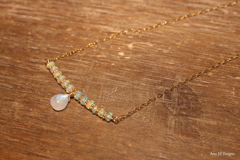 May include: A delicate gold necklace featuring a row of small opal beads and a teardrop-shaped moonstone pendant. The necklace is set against a wooden background, showcasing its elegant design.
